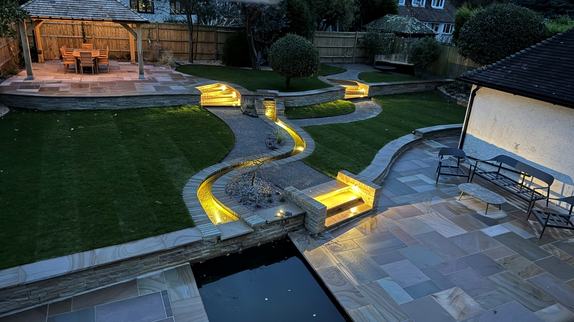 A garden landscape at twilight, featuring lit paving and a water feature